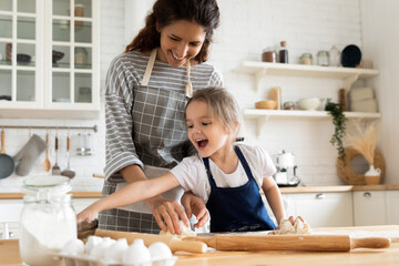 Close up mother playfully teaching little daughter to roll out dough standing at table in kitchen. Smiling caring mum and cute girl using rolling pin on wooden board, preparing dinner, cooking flour.