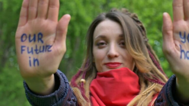 Modern serious girl with long dreadlocks is showing hands with written slogan 'Our future in your hands' on green tree background. Responsibility, climate change concepts