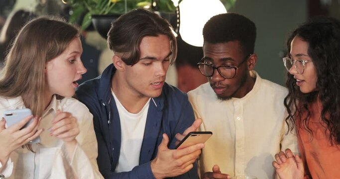 Close up view of young guy showing smartphone content to his friends and smiling. Cheerful millennial people using their phones while sitting in cafe while spending time together.