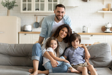Close up portrait picture of happy young family of four people sitting on couch at home. Smiling parents with little children hugging looking at camera posing for photo in living room.