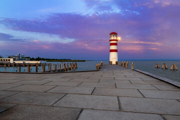 A pier leading to the lake and a red and white lighthouse at Lake Neusiedl in Podersdorf, Austria. In the background is a dramatic sky at sunrise.