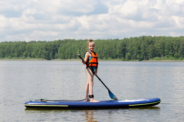 Sup surfing on the lake. Beautiful girl stands on a surfboard with a paddle in her hand. Orange vest. Summer day.