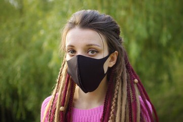 Modern girl with dreadlocks wearing textile mask on her face to protect herself from Coronavirus Covid-19 spending a time in the park outdoors