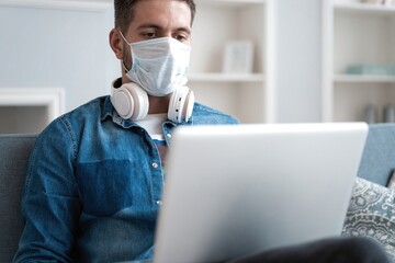 Fototapeta na wymiar Young caucasian man working from home, wearing protective mask, using laptop. Coronavirus pandemic, covid 19 quarantine