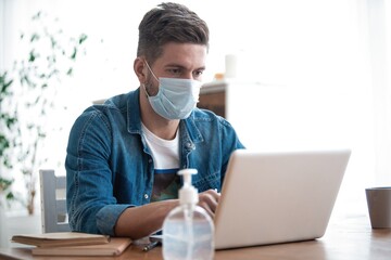 Young caucasian man working from home, wearing protective mask, using laptop. Coronavirus pandemic, covid 19 quarantine