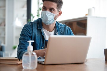 Young caucasian man working from home, wearing protective mask, using laptop. Coronavirus pandemic, covid 19 quarantine