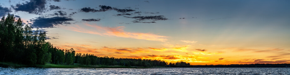 Obraz na płótnie Canvas Panorama of Scandinavian white night at polar and subpolar area in Sweden. Midnight Sun just below horizon line, sun rays illuminate highest clouds in atmosphere. Lake and forest at coast line