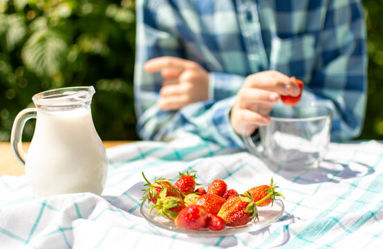 On A Wooden Table With A White Cloth In A Blue Check, There Is A Glass Pitcher Of Cream, Next To A Plate Of Strawberries, And In The Background A Child In A Checked Shirt