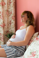 A smiling pregnant woman is sitting in bed, looking trough the window.