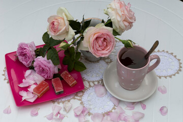 On a white round table on a lace napkin is a bouquet of pink roses, on a plate are two roses and marmalade. Nearby is a cup of tea..