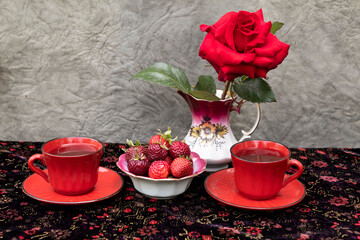 On a dark tablecloth are two cups of tea, a bowl of strawberries and a red rose in an elegant milk jug.