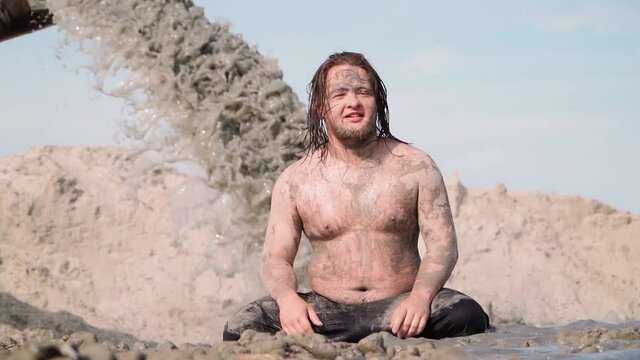 Funny Fat Man with Long Hair Sits in the Mud. Against the background pouring Black Dirty Water from the Pipe. Environmental pollution. Slow motion