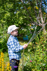 a child in a white cap and blue plaid shirt, watering the garden with a garden hose.
