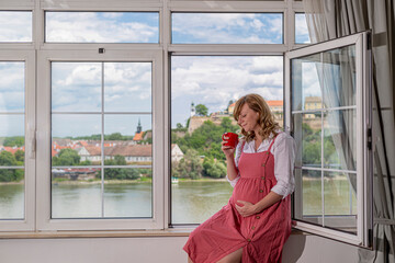 A pregnant caucasian woman sitting by the window with a cup of coffee or tea