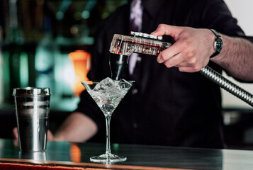 Bartender uses fountain soda gun to finish the martini style wine glass filled with ice, near a...