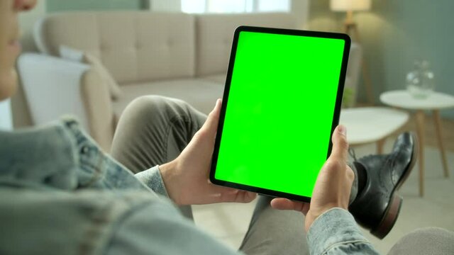 Over The Shoulder View Young Man at Home Sitting on a Chair Using With Green Mock-up Screen Tablet. Male Using Touchscreen Device, Browsing Internet. Concept of Green Screen and Chroma Key.