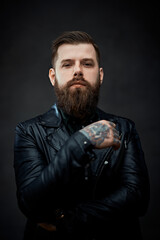 Cool bearded tattooed man in a leather jacket looking on the camera in a dark studio