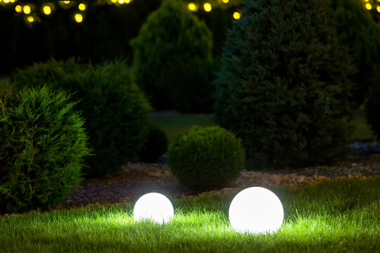backyard ground 2 light garden with lantern electric lamp with sphere diffuser in green grass with thuja bushes and stone mulching in park with landscaping, closeup night scene nobody.