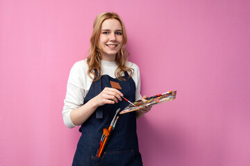 girl artist holds brushes and a palette and smiles on a pink background, student of art school,...