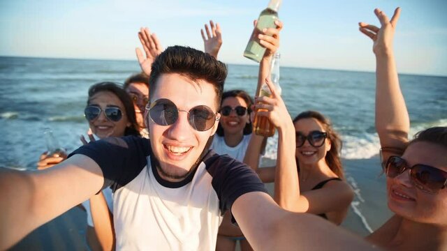 Pov view of young friends taking selfie having fun with drinks on sea beach on sunset. Online video call: man looking at smartphone camera on tropical island, women toasting lemonade, waving hands.