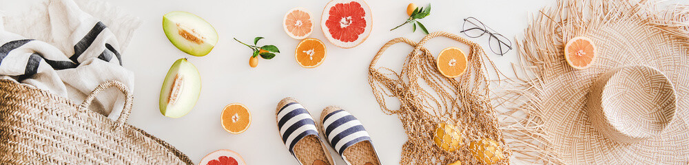 Summer mood layout. Flat-lay of summer natural espadrillas, straw sunhat, beach rafia and net bag, beach towel, sunglasses and fresh fruit over white plain background, top view, wide composition