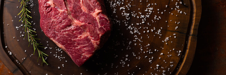 Top view of raw Chuck Eye beef steak seasoned with coarse salt and rosemary, on a dark wooden...