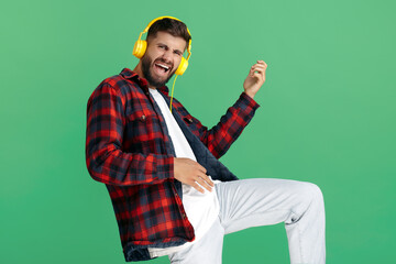 Joyful bearded hipster young man in plaid shirt listening music in headphones and dancing over green background.