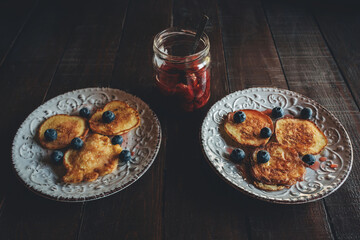 two white plates with pancakes, blueberry berries and jam on a dark wooden table side view