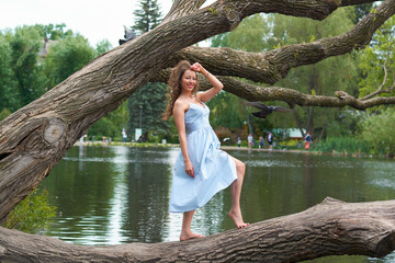 a beautiful curly haired girl in a blue dress stands on a large tree against the background of a Park with a lake