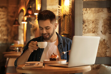 Young blogger with laptop eating dessert and drinking coffee at table in cafe