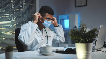 Medium shot of Young african doctor showing how to wear medical mask by video call on computer. He working home because of pandemic.