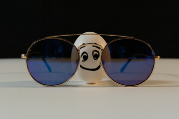Large White Chicken Egg with face drawn on wearing sunglasses with black background and white floor photograph funny