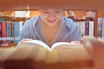 Young Asian woman university student reading book sitting by bookshelf in college library for education