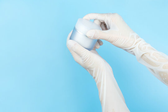 Hand in white latex medical gloves holding cream jar on blue background with copy space