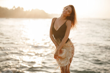 Naklejka premium Portrait of attractive blonde girl woman with long hair posing on rocky beach on sunset background, hold straw hat in her hands and look at the camera