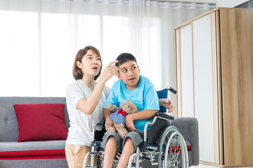 asian woman taking care disabled child on wheelchair in living room. both looking at something.