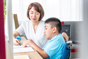 asian woman teaching disabled child on wheelchair with notebook in living room and blurred foreground.