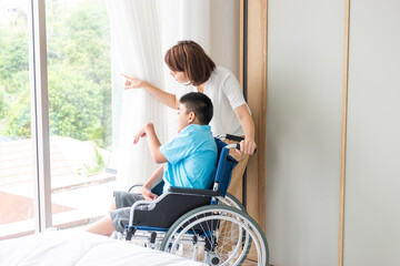 asian woman taking care disabled child on wheelchair in bedroom. both looking at something beside...