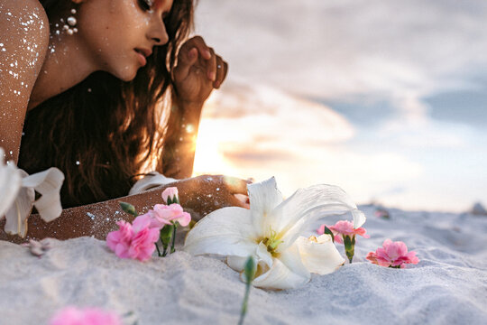 Pink lilies lying on sand foreground and young beatiful woman lying on sand at background on the beach at sunset
