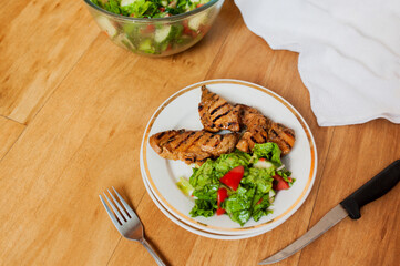 simple and healthy home-cooked meal in an ordinary home kitchen, grilled meat and salad with fresh summer vegetables, a silver fork and knife on a wooden table top with a cotton napkin