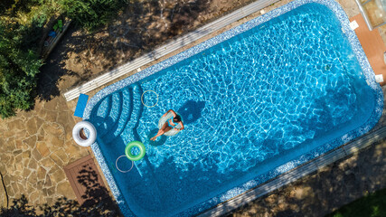 Aerial view of beautiful girl in swimming pool from above, swim on inflatable ring donut and has fun in water on family vacation on tropical holiday resort
