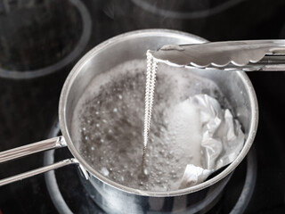 workshop for cleaning tarnished silver with aluminum foil and baking soda - tongs removes clean silver chain from boiling solution of baking soda with aluminum foil in pot on electric stove