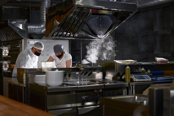 Fototapeta na wymiar mask on the chef's face in a restaurant: cooks in an open kitchen, a client sees work during a pandemic