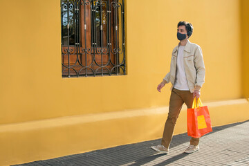 nueva normalidad coronavirus, joven con barbijo de cuidado está caminando en la vereda con una bolsa de compras, sobre pared color amarillo 