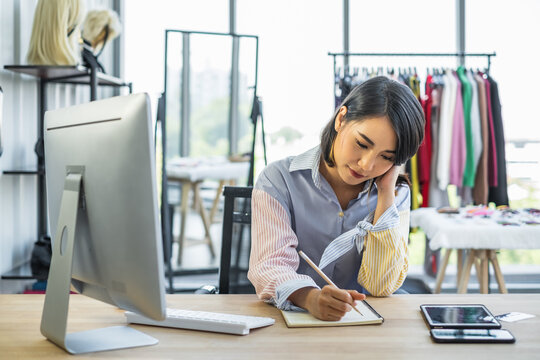 Young Asian Girl Fashion Shop Owner Siting In Front Of A Computer Writing On A Notebook Inside A Clothing Store.