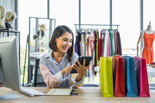 Young Asian woman sitting in front of a computer in a clothing fashion shop smiling looking at tablet with multicolor shopping bags on the desk.