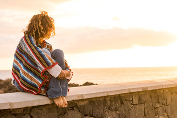 Unrecognizable lonely woman sit down on a wall looking and enjoying the sunset on the sea - coloured trendy mexican poncho style and people in outdoor leisure relax activity