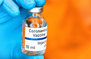 Hand in blue medical gloves holding small glass vial with label Coronavirus vaccine (sticker own design, not real product), blurred bright orange background
