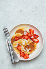 Traditional breakfast pancakes with strawberries and coconut chips in white plate, light background, top view.