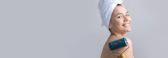 Brunette with a towel on her head using roller for face massage isolated on grey background